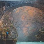 Mies seisoo joen reunalla suuren kivisillan alla syksyisessä maisemassa.