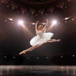 Valkoasuinen tanssija hyppäämässä korkealle lavalla.