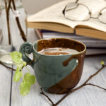 Kahvikupin takana häämöttää avoin kirja ja silmälasit.