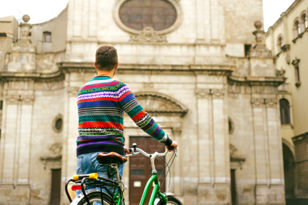 Värikkääseen neulepaitaan pukeutunut mies pyörän kanssa katsoo vanhaa rakennusta.