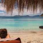 Nainen istuu aurinkotuolissa olkivarjon alla rannalla.