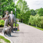 Vehreän puiston kivinen kävelykatu, jonka reunalla miehen, naisen ja koiran patsaat.