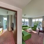 kaksi huonetta, jossa toisessa näkyy vähän sänkyä ja tv, toisessa kaksi vihreää sohvaa ja nojatuoli, sohvapöytä ja kirjoituspöytä