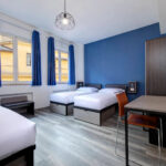 Hotellihuone jossa siniset verhot ja seinä, kirjoituspöytä ja kolme vuodetta